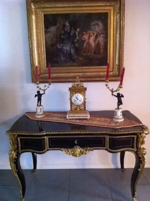 Une table ornée de touches dorées accueille une horloge détaillée et deux bougeoirs. Au-dessus, un tableau encadré représentant un personnage historique avec des enfants est accroché au centre du mur. Cette élégante disposition témoigne d'une consommation raffinée, complétée par un chemin de table à motifs sous les objets.