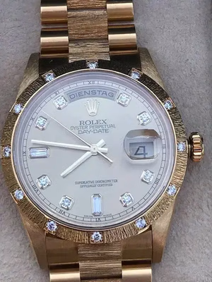 Une montre Rolex Oyster Perpetual Day-Date en or, que j'adore pour son cadran blanc et ses index sertis de diamants. Le guichet de date affiche « 4 » et le jour en allemand, « DIENSTAG ». La lunette est ornée de diamants scintillants.