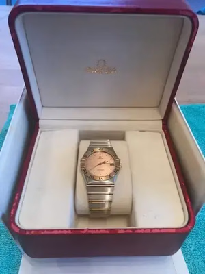 Une montre-bracelet en or au cadran rose repose dans un coffret Omega rouge ouvert, invitant à l'achat. L'intérieur du coffret est de couleur crème, avec le logo Omega sur le couvercle. La montre, dotée d'un bracelet en métal et de chiffres romains sur la lunette, est prête à être achetée par son prochain propriétaire.
