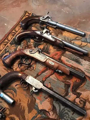 Une collection de quatre pistolets anciens est exposée sur un tissu aux motifs complexes, incarnant « Ce que j'achète ». Les manches en bois et les détails métalliques ornés varient en style, avec des marteaux et des détentes visibles, reposant sur un tissu riche et naturel.