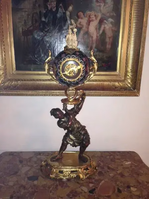 Un calice antique décoratif, orné de détails dorés et d'une base sombre, sur lequel un personnage le soutient, attire le regard comme un trésor que je pourrais acheter lors d'une journée de « ce que j'achète ». Derrière lui, un tableau encadré représentant des personnages classiques est accroché au mur, sur la surface en marbre.