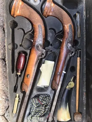 Un coffret en bois contenant deux pistolets anciens aux poignées ornées est disponible à l'achat. L'ensemble comprend un tournevis à manche en bois, une poire à poudre, des baguettes et d'autres accessoires. L'intérieur patiné du coffret ouvert révèle un charme exquis.