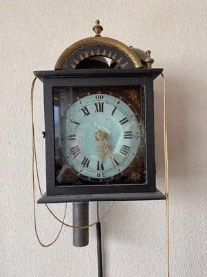 Horloge murale ancienne avec cadran à chiffres romains et aiguilles ornées, encadrée dans un cadre noir rectangulaire. Des touches dorées décoratives ornent le dessus. Une cordelette ou un fil suspendu de chaque côté invite à l'achat de cette pièce intemporelle. L'horloge est fixée sur un mur clair.