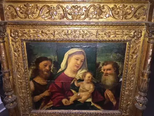 Un tableau orné, encadré d'or, représente une femme vêtue d'une robe rouge et blanche tenant un enfant, entourée de deux hommes barbus vêtus de robes sombres. Ce magnifique tableau évoque la beauté sereine du passé, avec en arrière-plan un paysage arboré.