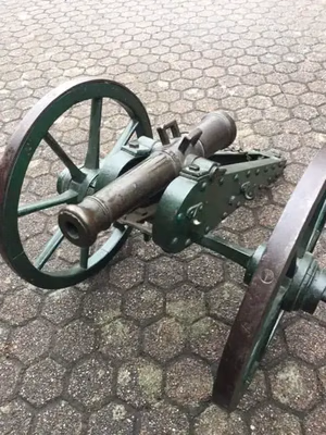 Exposé sur une surface pavée, ce petit canon ancien, idéal pour les amateurs de vintage, est doté de roues à rayons en bois et d'un corps en métal patiné vert. Son canon légèrement pointé vers le haut ajoutera du charme à votre collection de produits uniques.