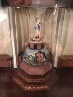 Une vitrine vitrée présente une pièce en céramique ornée sur un socle hexagonal en bois. L'œuvre est ornée d'un dôme peint représentant des scènes extérieures et mythologiques, surmonté d'une petite statue d'un personnage en costume.