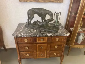 J'achète un magnifique meuble en bois avec un dessus en marbre, présentant des tiroirs ornés et des poignées en laiton. Une sculpture en bronze de deux lévriers et un petit vase y sont exposés. À droite, une vitrine avec des objets décoratifs est partiellement visible.