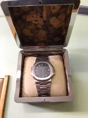 Une montre-bracelet en métal argenté au cadran foncé, présentée dans un boîtier en bois matelassé ouvert avec un intérieur à motif liège. La montre est soigneusement placée et le boîtier repose élégamment sur une surface claire.