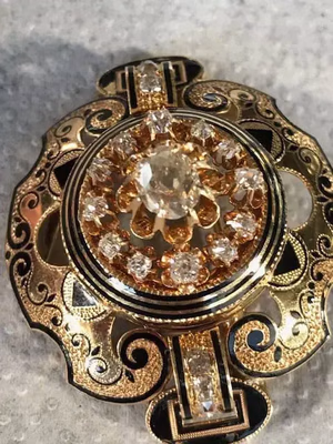 Broche complexe en or et noir ornée d'une pierre ovale centrale entourée de pierres plus petites. Son design détaillé, composé de motifs volutes, crée un effet ornemental et élégant sur un fond gris texturé. Une pièce incontournable pour son élégance intemporelle.