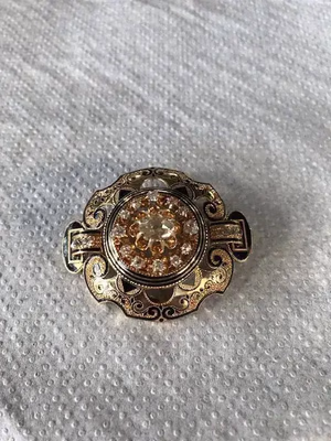 Une broche circulaire décorative avec des détails en filigrane ornés, comportant une pierre précieuse centrale transparente entourée de pierres d'ambre plus petites, repose sur une surface blanche texturée - un véritable témoignage de ce que j'achète pour une élégance intemporelle.