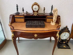 Un bureau en bois orné, surmonté d'une horloge vintage et de petites figurines, incarne l'essence même de « ce que j'achète ». Des livres à dos décoratif bordent les étagères, tandis qu'un grand livre fermé en constitue le centre. Une autre horloge est gracieusement posée au sol à côté.