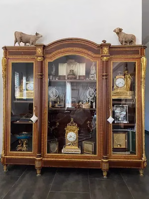 Une vitrine en bois ornée de portes vitrées présente une variété d'horloges et de figurines décoratives, parfaites pour ajouter une touche d'élégance à votre liste de courses. Deux statues d'animaux trônent au sommet, tandis que de délicats détails dorés rehaussent son charme, le tout se dressant fièrement sur un sol carrelé sombre.