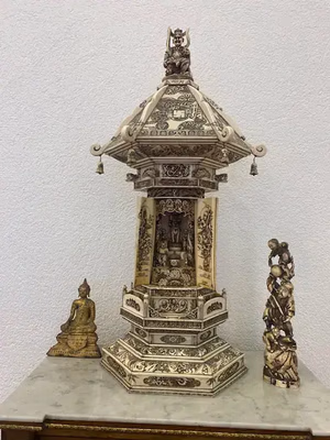 Tour d'ivoire ornée de sculptures complexes, surmontée d'une petite statue. Un Bouddha assis à gauche et un délicat groupe de personnages sculptés à droite, le tout posé sur une table en marbre, adossé à un mur blanc. Une pièce charmante à acquérir pour votre collection de produits raffinés.