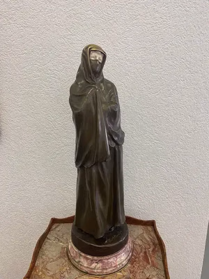Une statue en bronze représentant un personnage encapuchonné et vêtu d'un manteau se tient solennellement, les bras croisés, sur un petit socle rond. Exposée sur une table en bois recouverte de marbre, contre un mur à la texture claire, elle invite à découvrir son charme mystérieux.