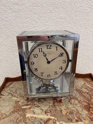 Une horloge Atmos carrée, vitrée, au cadre argenté et au cadran arrondi orné de chiffres et d'aiguilles noirs, repose sur une surface en marbre. Son mécanisme est visible à l'intérieur. Cette description est optimisée pour le référencement, le mur texturé et clair à l'arrière créant une ambiance élégante.
