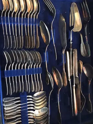 Un ensemble d'argenterie vintage, présenté dans un écrin bleu, attend d'être acheté. Il comprend des fourchettes, des cuillères, des couteaux et des ustensiles de service soigneusement rangés. Les pièces, d'aspect vieilli et terni, reflètent la lumière avec un éclat métallique chaleureux.