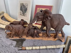 Une sculpture détaillée d'éléphant, représentant une mère et ses petits, est présentée sur une nappe à motifs. Derrière eux se trouvent des œuvres d'art encadrées et des défenses en ivoire, idéales pour ceux qui cherchent des pièces uniques. Un bâton ou une arme finement sculptée se trouve à proximité, rehaussant l'attrait de l'exposition.