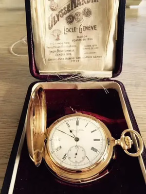 Montre de poche vintage Ulysse Nardin à chiffres romains, logée dans un boîtier ouvert doublé de velours rouge. Le couvercle arbore l'étiquette de la marque, qui précise ses origines au Locle, à Genève. Affichée sur une surface en bois, cette pièce intemporelle promet aux collectionneurs du monde entier un « j'achète ».