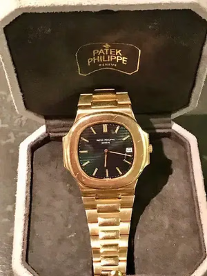 Une montre Patek Philippe en or au cadran sombre est élégamment présentée dans un écrin ouvert gris et noir, mettant en valeur le logo emblématique de la marque sur le couvercle. C'est vraiment ce que j'achète pour s'offrir une sophistication intemporelle.