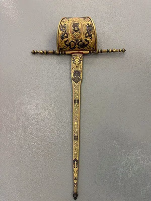 Une épée ancienne et ornée, ornée de détails complexes dorés et noirs sur la poignée et la lame. Son design, orné de motifs floraux et d'un cimier, est idéal pour les collectionneurs ou ceux qui cherchent des pièces uniques. Présentée verticalement sur un fond gris uni, c'est un objet remarquable.