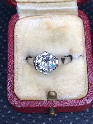 Une bague en argent ornée d'un gros diamant rond repose dans un luxueux écrin couleur crème, dont la doublure en velours enveloppe la pierre précieuse. Ouvert et accueillant, le coffret arbore un rouge profond et un petit fermoir métallique : une élégance intemporelle.