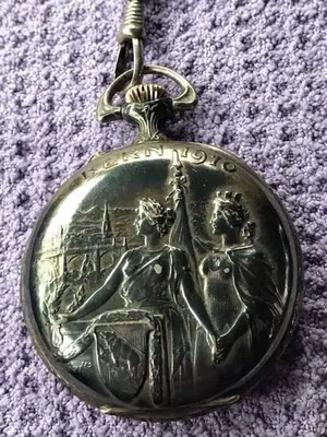 Montre de poche ancienne en argent ornée de gravures complexes représentant deux figures royales, des boucliers et des drapeaux. L'inscription « Bern 1910 » est gravée sur le dessus. Sur fond de tissu violet texturé, c'est une pièce incontournable pour son élégance intemporelle et son charme historique.
