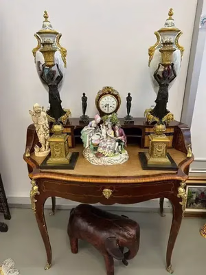 Une table vintage en bois aux détails dorés orne deux lampes décoratives, une horloge et une figurine en porcelaine. Une petite sculpture d'éléphant trône au sol en dessous. J'adore cette présentation soignée et élégante, avec un tableau encadré adossé au mur en arrière-plan.
