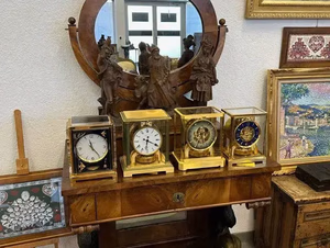 Une exposition de quatre horloges ornées sur une table en bois sculpté, avec un miroir circulaire et des sculptures décoratives, invite à la découverte d'une décoration unique. En arrière-plan, des œuvres d'art encadrées et des broderies ornent un mur blanc immaculé.