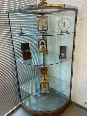 Une vitrine en verre incurvé est élégamment placée dans un coin de la pièce, présentant plusieurs horloges et garde-temps vintage sur ses trois étagères. Idéale pour ceux qui cherchent des pièces de décoration uniques, l'arrière-plan est orné de stores verticaux qui masquent partiellement une fenêtre.