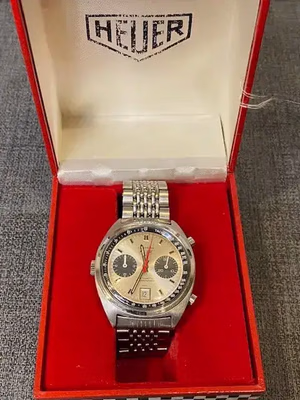 Montre chronographe Heuer vintage avec bracelet en acier inoxydable, présentée dans un coffret ouvert rouge et blanc. Idéale pour les connaisseurs, elle est dotée d'un cadran beige, de deux compteurs et d'un guichet de date. Le couvercle du coffret arbore le logo emblématique Heuer.