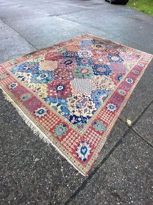 Un tapis persan coloré, aux motifs floraux et géométriques complexes, est posé sur un trottoir. Que ce soit dans des tons rouges, bleus ou autres couleurs vives, il séduit par son design et sa frange sur un bord. La rue est légèrement humide, suggérant une pluie récente ; un décor idéal pour acheter cette pièce exquise.