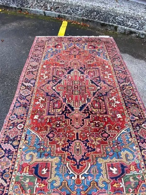 Un tapis oriental aux riches motifs géométriques et floraux complexes, aux tons rouge, bleu et crème, est posé sur un trottoir extérieur. Son imposant motif en losanges au centre invite tous les passants à s'arrêter, à admirer sa beauté et à se dire « j'achète » en l'imaginant chez eux.