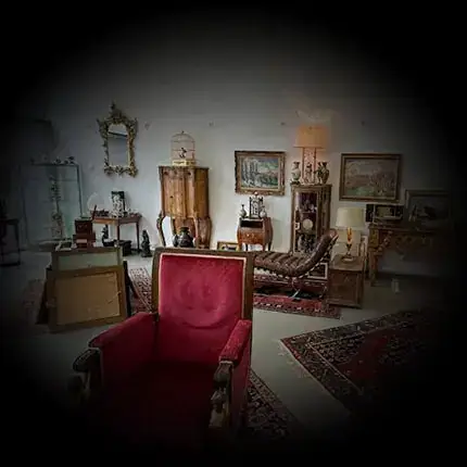 Une pièce tamisée, décorée de meubles et d'objets vintage. Un fauteuil rouge trône au premier plan, entouré de tapis ornés, de tableaux aux murs et d'armoires anciennes. Un éclairage chaleureux confère à l'espace une ambiance chaleureuse et nostalgique.