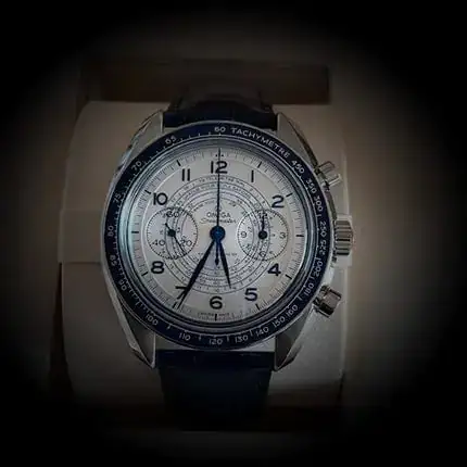 Montre-bracelet de luxe avec cadran blanc et lunette bleue. Elle est dotée de trois sous-cadrans et de chiffres arabes. Elle est dotée d'un bracelet en cuir bleu et est présentée dans un écrin.