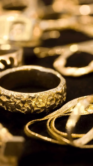 Gros plan sur diverses bagues et chaînes en or sur une surface sombre. L'accent est mis sur une bague en or texturée, entourée de bijoux flous et brillants, créant un effet luxueux et élégant.