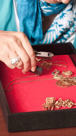Une personne portant un foulard bleu utilise une pince pour réparer un collier en or sur une surface rouge. Divers bijoux en or sont éparpillés un peu partout. Une bague à motif géométrique est portée à son doigt.