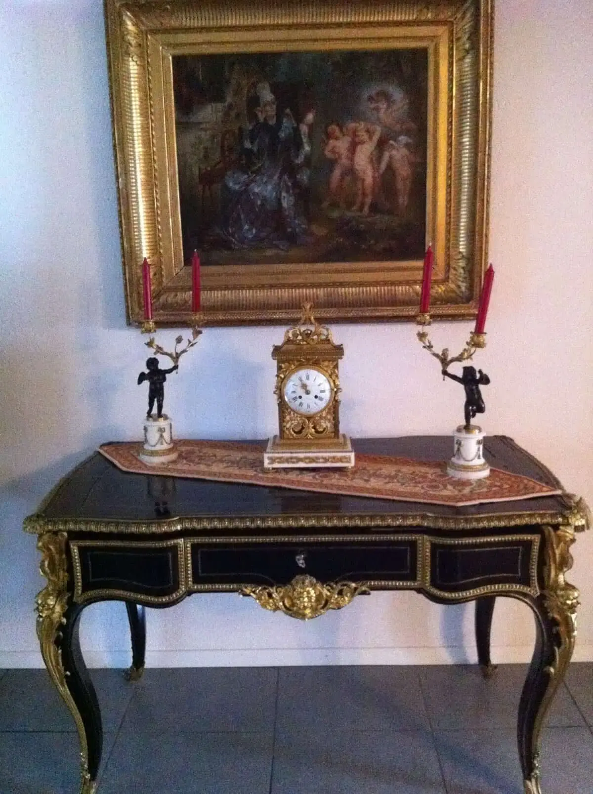 Une table noire et or ornée de sculptures complexes, évoquant l'élégance de Genève, est ornée d'une horloge ancienne centrale. Deux bougeoirs ornés de bougies rouges sont disposés de chaque côté. Un tableau détaillé, encadré d'or, est accroché au mur au-dessus de la table.