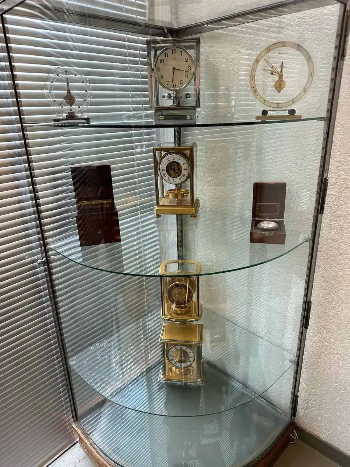 Une vitrine en verre présente des horloges vintage de Genève sur deux étagères. L'étagère supérieure accueille trois petits trésors, tandis que celle du milieu présente trois pièces plus grandes et aux motifs complexes. La vitrine, adossée à un mur et dotée d'une fenêtre et de stores, incarne une élégance intemporelle.