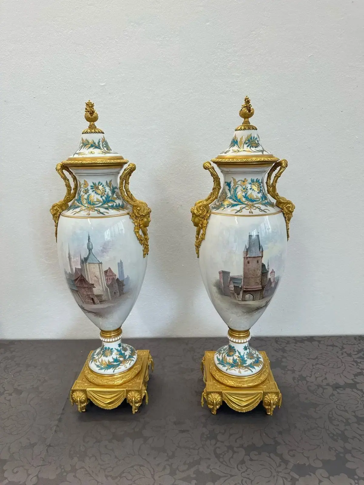 Deux vases en porcelaine ornés de détails dorés reposent sur une surface à motifs rappelant le charme pittoresque de Vevey. Chaque vase présente des scènes peintes complexes de bâtiments et de paysages, surmontées de fleurons décoratifs. Leurs bases sont ornées d'ornements dorés.