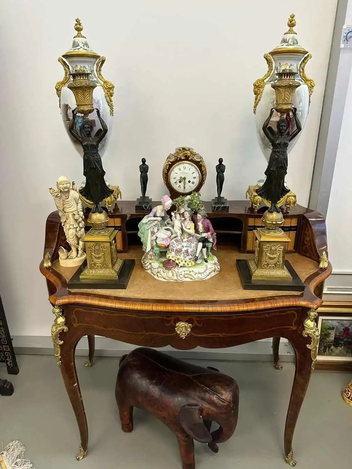 Une table en bois ornée aux pieds incurvés, rappelant l'élégance charmante de Vevey, accueille deux grandes urnes décoratives, une petite horloge et une figurine de personnages du XVIIIe siècle. Une petite sculpture d'éléphant repose au sol sous la table.