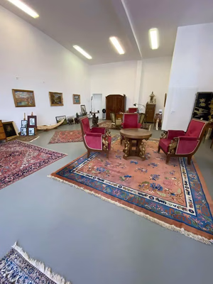 Une pièce spacieuse présente une variété de tapis au sol, avec des sièges roses et dorés sur l'un d'eux. Les murs sont ornés de tableaux encadrés, tandis que des meubles anciens invitent à l'admiration. On pourrait facilement être tenté d'acquérir une décoration aussi raffinée.