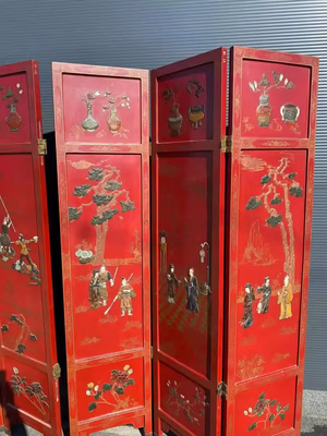 Un ensemble de paravents rouges richement décorés, représentant des scènes peintes complexes représentant des personnages, des arbres et des vases dans un style artistique asiatique. Le fond est un mur sombre et strié, précisément ce que j'achète pour une touche d'élégance.