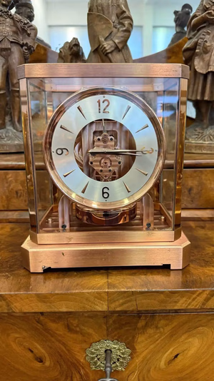 Une horloge de cheminée vintage Sion en verre et cuivre repose sur une surface en bois poli, son mécanisme complexe étant visible à l'intérieur. Des figurines en bronze sont exposées en arrière-plan.