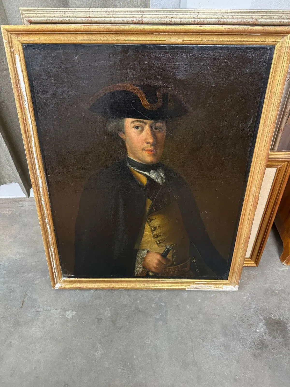 Une peinture à l'huile encadrée représentant un homme en tenue historique, portant un chapeau tricorne et un manteau sombres, tenant un document ou un livre, se dresse sur un sol en béton - une scène rappelant Neuchâtel au XVIIIe siècle.