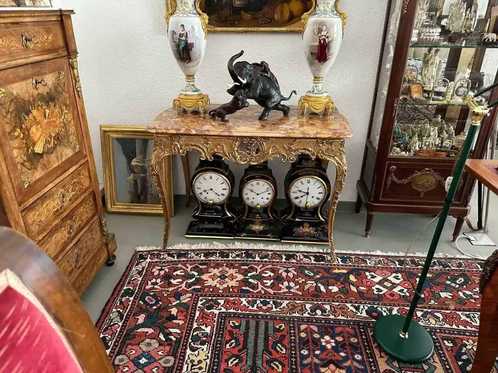 Cette pièce remplie d'antiquités, avec sa table ornée d'un plateau en marbre et ses objets décoratifs, dont une figurine d'éléphant noir, est l'endroit idéal pour des achats uniques. En contrebas, trois horloges sont posées sur un tapis aux motifs riches, entourées de meubles en bois et de vitrines en verre.