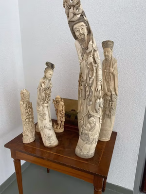 Un groupe de sculptures en ivoire finement sculptées est exposé sur une table en bois devant un mur blanc. Ces exquises figures, représentant des formes et des expressions humaines détaillées, séduisent les amateurs d'art qui souhaitent enrichir leur collection de pièces uniques tout en achetant des objets remarquables.