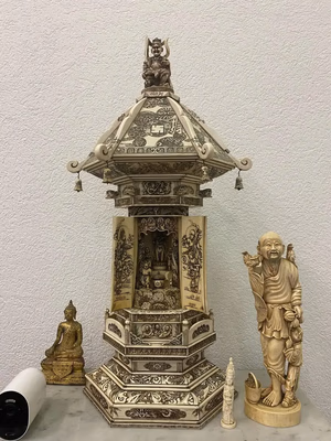 Un sanctuaire hexagonal orné de sculptures complexes abrite une petite divinité. Une figure est assise au sommet, entourée de deux statues : un Bouddha assis et un personnage souriant avec un bâton. À proximité repose un petit objet cylindrique. Parfait pour ceux qui recherchent une décoration unique – achetez cette pièce exquise dès maintenant !.
