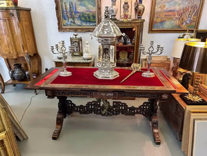 Une table en bois ornée, recouverte d'un plateau en velours rouge, est ornée d'une pièce décorative raffinée et de deux candélabres en argent. Autour de la table, divers tableaux encadrés et des meubles anciens, dans une pièce richement décorée, créent une ambiance captivante que l'on aimerait bien s'approprier pour son propre intérieur.