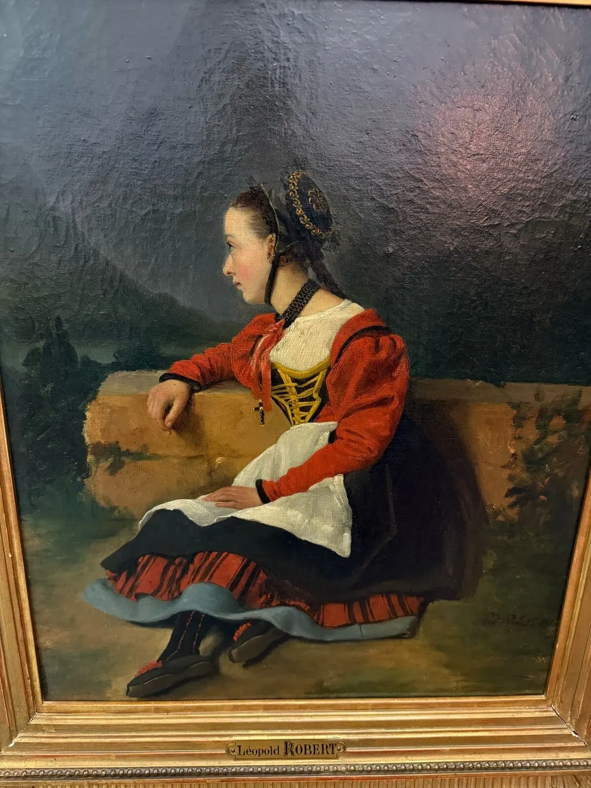 Une jeune femme en costume traditionnel européen bernois, avec veste rouge, tablier blanc, jupe rayée et coiffe, est assise de profil sur un banc de pierre, sur fond sombre et naturel. Le tableau est encadré et signé.