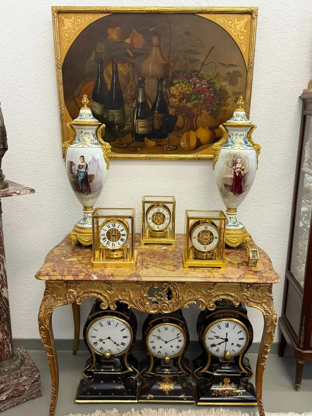 Une table décorative de Bâle présente des horloges ouvragées et deux grands vases ornés de personnages peints, sous une nature morte encadrée représentant des fruits et des bouteilles de vin. Le plateau en marbre est orné de détails dorés complexes.